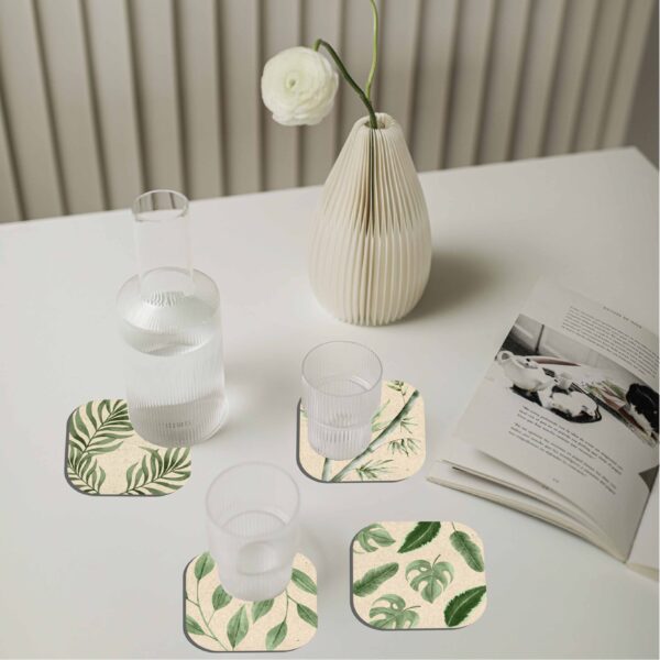 Portavasos ecológicos con diseños de hojas tropicales sobre mesa blanca, acompañados de vasos de vidrio, florero decorativo y revista abierta. Producto de la subcategoría Línea Ecológica dentro de regalos corporativos, ideal para espacios sostenibles y modernos.